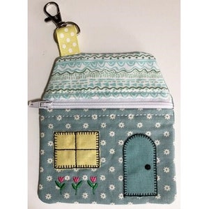 May include: A house-shaped coin purse with a zipper closure. The purse is light blue with a floral pattern and features a yellow window, a blue door, and embroidered flowers. A yellow and white polka dot strap is attached to a silver clasp.