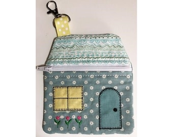 May include: A house-shaped coin purse with a zipper closure. The purse is light blue with a floral pattern and features a yellow window, a blue door, and embroidered flowers. A yellow and white polka dot strap is attached to a silver clasp.