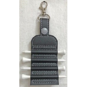 Pode incluir: Um porta-tees de golfe de couro preto com um clipe prateado e cinco tees de golfe brancos. O porta-tees tem um design costurado e um botão cinza.