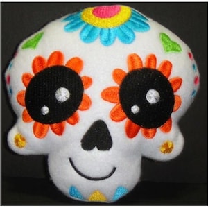 May include: A white plush skull with embroidered details. The skull has orange, blue, yellow, and green flowers, black eyes, and a black heart-shaped nose. The skull is smiling.