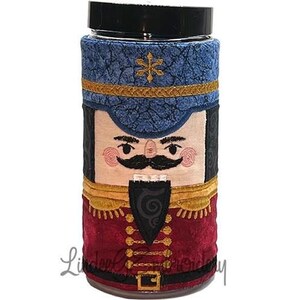 Può includere: Un viso di schiaccianoci rosso, oro e blu su un barattolo di vetro con un coperchio nero. Lo schiaccianoci indossa un cappello blu con un bordo dorato e un'uniforme rosso e oro.