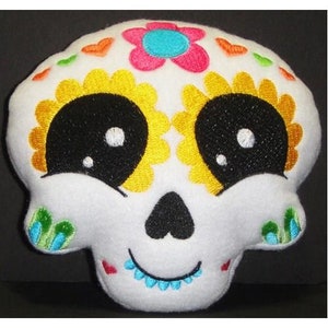 May include: White plush skull with embroidered details. The skull has black eyes with white highlights, a yellow flower design, and a pink flower with a blue centre. The skull is smiling with a blue and white trim around the mouth.