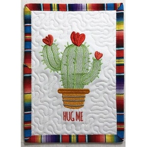 May include: A white quilted coaster with a colorful striped border. The coaster features an embroidered cactus with red flowers and the text "HUG ME".