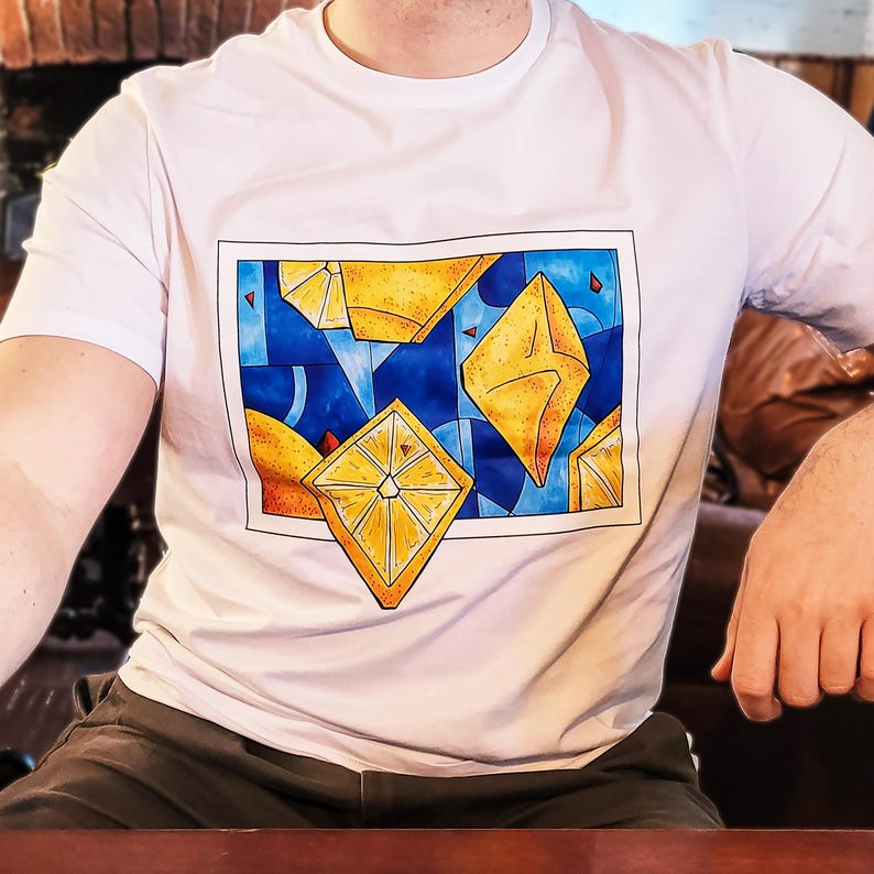 May include: White t-shirt featuring a geometric design with yellow and orange diamond shapes set against a blue background. The design is framed by a black border. The shirt is a classic crew neck style.