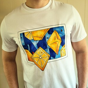 May include: White t-shirt featuring a geometric design of yellow and orange shapes against a blue background. The design is framed with a black border. The shirt is made of a soft, comfortable material.