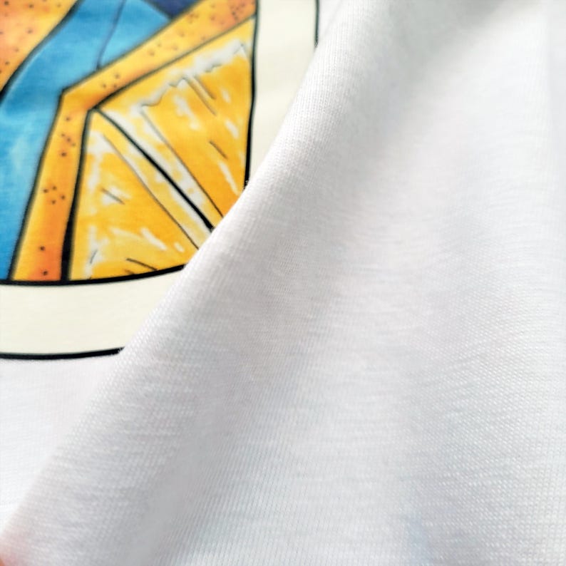 May include: White t-shirt with a colorful graphic print. The graphic features a geometric design with yellow, orange, and blue elements. The fabric appears soft and lightweight, with visible texture.