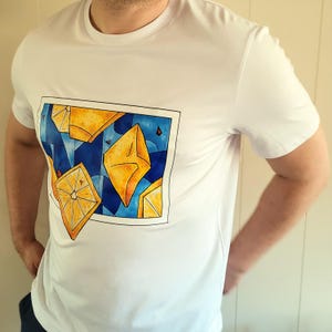 May include: White t-shirt featuring a rectangular graphic with yellow geometric shapes and lemon slices against a blue background. The shirt is a classic crew neck style.