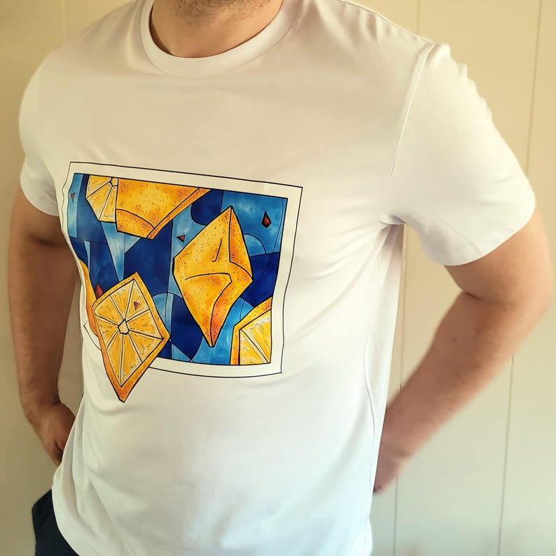 May include: White t-shirt featuring a rectangular graphic with yellow geometric shapes and lemon slices against a blue background. The shirt is a classic crew neck style.