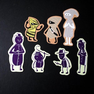 Puede incluir: Una colección de pegatinas de dibujos animados con varios personajes. Las pegatinas incluyen figuras que se asemejan a momias e individuos con sombreros fedora y gabardinas, con una combinación de colores púrpura y blanco. Sobre un fondo negro.