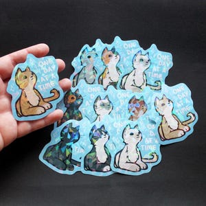 May include: A collection of cat stickers with a holographic effect. Each sticker features a stylized cat in various colors and patterns, with the text "ONE DAY AT A TIME" on a light blue background.