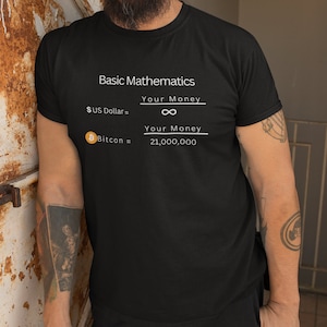 Puede incluir: Una camiseta negra con texto blanco que dice "Basic Mathematics" y tiene dos ecuaciones: "$US Dollar = Your Money ∞" y "฿ Bitcoin = Your Money 21,000,000".