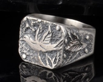 May include: A silver ring with a square face featuring an engraved design of a bird in flight surrounded by leaves.