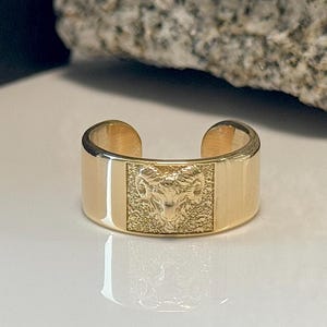 May include: A gold ring with an open design. The ring has a rectangular centre with a detailed engraving of a ram's head. The ring is set against a white surface with a rock in the background.