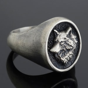 May include: A silver ring featuring a wolf head design. The ring is oval shaped and has a textured finish.
