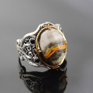 Könnte beinhalten: Ein silberner Ring mit einem verzierten Design und einem großen ovalen Edelstein. Der Edelstein ist eine Mischung aus weißen, braunen, gelben und schwarzen Farben.