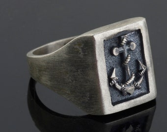 Könnte beinhalten: Ein silberner Ring mit einer quadratischen schwarzen Einlage, die ein detailliertes silbernes Ankerdesign zeigt.