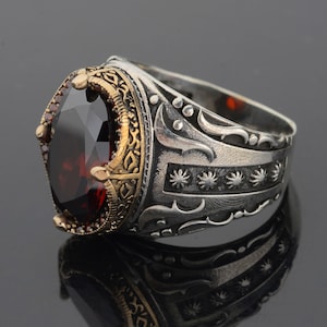 Könnte beinhalten: Ein silberner Ring mit einem verzierten Design und einem großen, roten Edelstein. Der Ring hat eine goldfarbene Band und komplizierte Details.