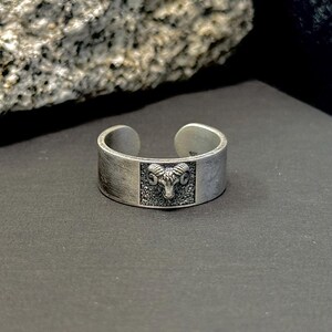 May include: A silver ring with an open design. The ring has a rectangular textured panel with a detailed ram's head. The ring is set against a dark background with a rock in the upper left corner. The ring is likely made of silver.