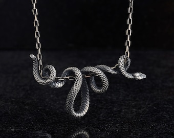Puede incluir: Un collar de serpiente de plata con un colgante de serpiente enroscada. La serpiente está detallada con escamas y tiene un aspecto realista.