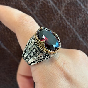 May include: A silver ring with a large, oval-shaped, dark red gemstone set in a gold-toned, ornate setting. The ring has intricate details and a textured finish.
