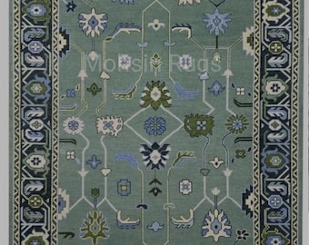 May include: A rectangular area rug featuring a sage green background with a central symmetrical pattern of stylized floral and geometric motifs in shades of blue, white, and olive green. The border is dark blue with intricate white patterns.