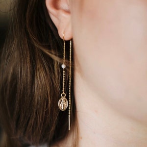 Puede incluir: Pendientes de cadena de oro con un pequeño encanto de perla y un encanto de oro con un diseño religioso.