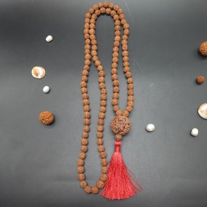 May include: A brown wooden mala necklace with a tassel. The necklace is made of 108 beads and has a large, round bead at the end. The tassel is red and is made of a soft, silky material.