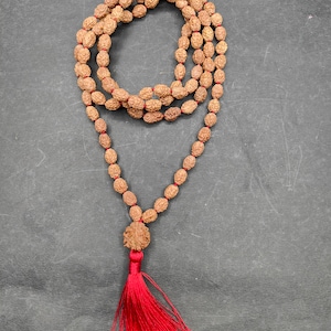 Può includere: Una collana di mala in legno marrone con un fiocco rosso. La collana è composta da 108 perle ed è usata per la meditazione e la preghiera.