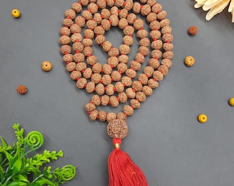 May include: A brown mala bead necklace with a red tassel. The beads are made of natural materials and are strung on a red thread. The tassel is made of red fabric and is tied to the end of the necklace.
