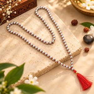 Puede incluir: Un collar largo que presenta una hebra continua de cuentas esféricas de tono plateado pulido ensartadas en un cordón rojo vibrante. El collar está dispuesto en un bucle doble y termina en una borla roja completa en el extremo.