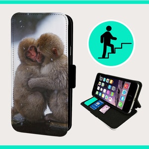 Puede incluir: Una funda de teléfono de cuero negro con una solapa que se pliega sobre la pantalla. La funda tiene una imagen impresa de dos monos abrazándose. Los monos son marrones y blancos y están en un entorno nevado.