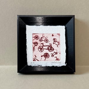 Puede incluir: Una obra de arte enmarcada con un patrón repetido de ilustraciones de bicicletas rojas sobre un fondo blanco. La obra está presentada en un marco negro. Las iniciales del artista "E.A." y la fecha "Natale/24" son visibles.