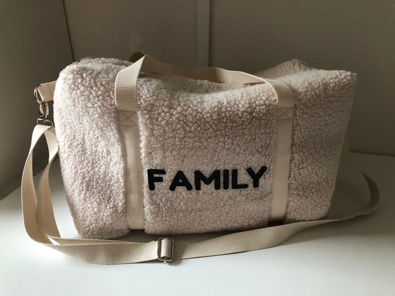 May include: A white faux fur duffel bag with a black strap and the word "FAMILY" embroidered on the front.