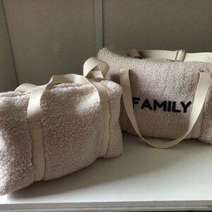 May include: Two white sherpa fabric duffel bags with white straps. The larger bag has the word "FAMILY" embroidered in black on the front.