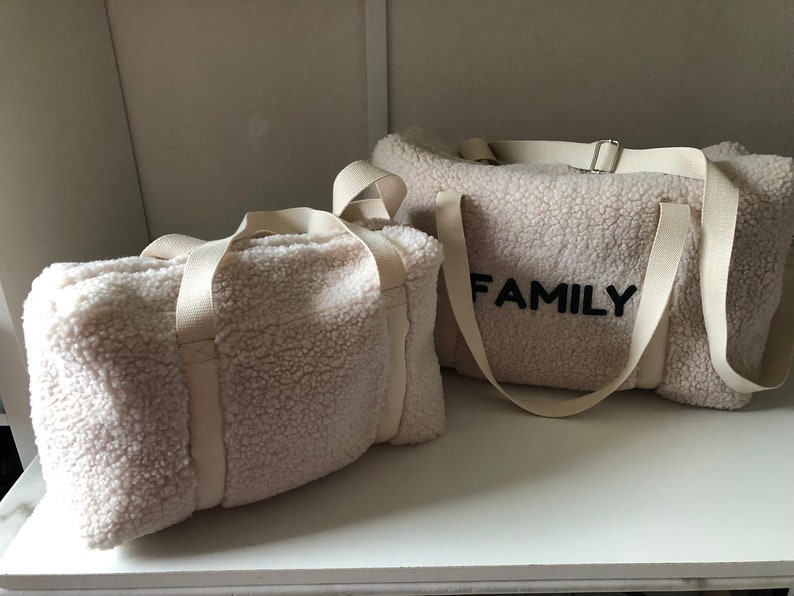 May include: Two white sherpa fabric duffel bags with white straps. The larger bag has the word "FAMILY" embroidered in black on the front.