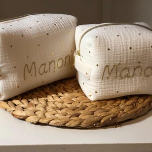 以下が含まれることがあります： 金色のグリッターと「マノン」と「ミアンオン」という名前が金糸で刺繍された、白い布製のジッパー付きポーチが2つ。ポーチは、編まれたわら製のマットの上に置かれています。