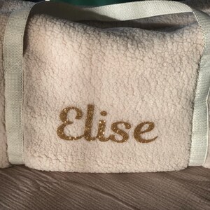 May include: White faux fur duffel bag with white straps and the name "Elise" embroidered in gold glitter.