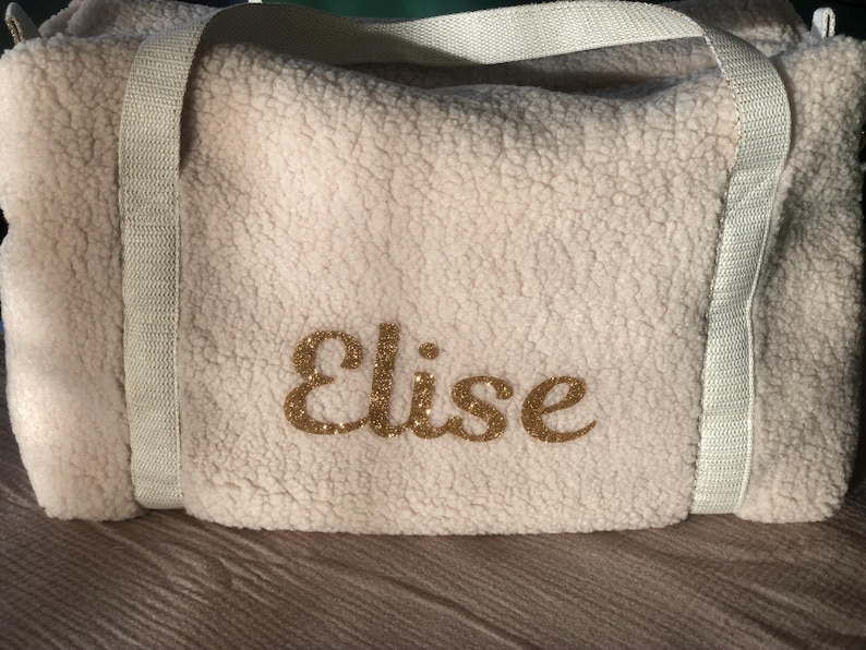 May include: White faux fur duffel bag with white straps and the name "Elise" embroidered in gold glitter.