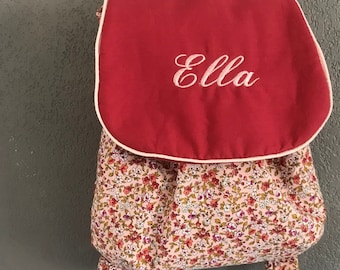 Op de afbeelding: Een kleine rugzak met een bloemenpatroon van roze, rode en witte bloemen op een lichte achtergrond. De bovenste flap is effen rood met lichtroze biezen en de naam Ella is in witte schrijfletters op de voorkant geborduurd.