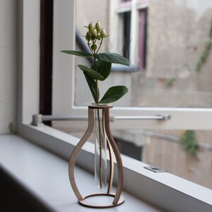 May include: A wooden vase with a clear glass test tube insert, holding a single stem of white flowers with green leaves. The vase is sitting on a white windowsill.