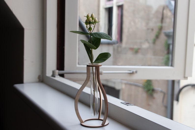 May include: A wooden vase with a clear glass test tube insert, holding a single stem of white flowers with green leaves. The vase is sitting on a white windowsill.