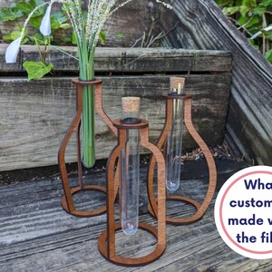 May include: Three wooden test tube holders with glass test tubes inside. The holders are brown and have a rounded shape. The test tubes are clear and are filled with green stems and white flowers.