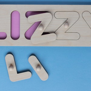 Op de afbeelding: Houten naam puzzel met de naam "LIZZY" met roze achtergrond. De letters zijn uitneembaar, met kleine houten knoppen voor eenvoudige bediening. De puzzel ligt op een blauw oppervlak.