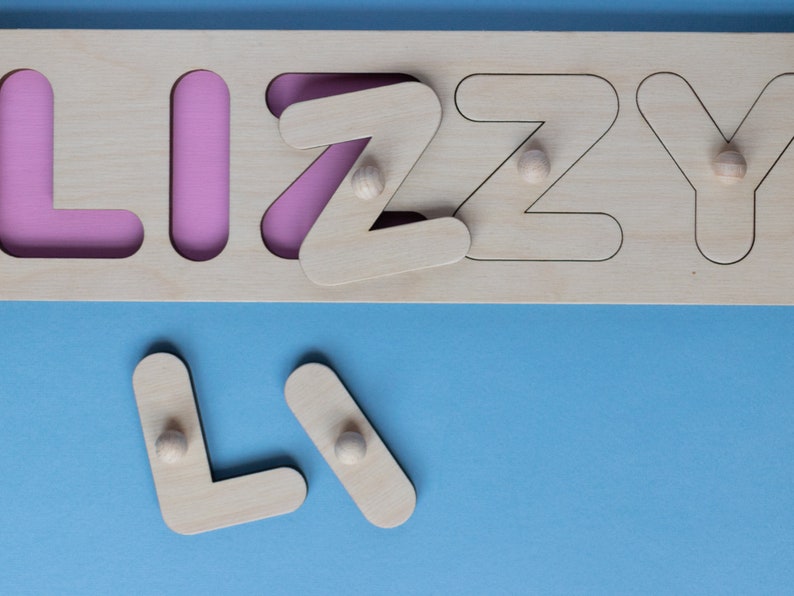 Op de afbeelding: Houten naam puzzel met de naam "LIZZY" met roze achtergrond. De letters zijn uitneembaar, met kleine houten knoppen voor eenvoudige bediening. De puzzel ligt op een blauw oppervlak.