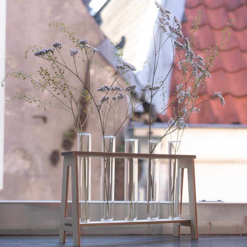 Puede incluir: Un soporte de madera blanco con seis tubos de ensayo de vidrio transparente que contienen ramitas de flores blancas.