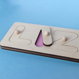 Op de afbeelding: Houten naam puzzel met de naam "LIZZ" met een lichtroze achtergrond voor de letter "A". De puzzel heeft een natuurlijke houten afwerking met afgeronde houten pinnen voor elke letter. De letters zijn uitgesneden en de puzzel ligt op een blauwe achtergrond.