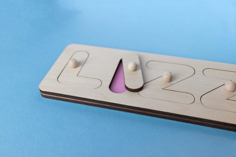 Op de afbeelding: Houten naam puzzel met de naam "LIZZ" met een lichtroze achtergrond voor de letter "A". De puzzel heeft een natuurlijke houten afwerking met afgeronde houten pinnen voor elke letter. De letters zijn uitgesneden en de puzzel ligt op een blauwe achtergrond.