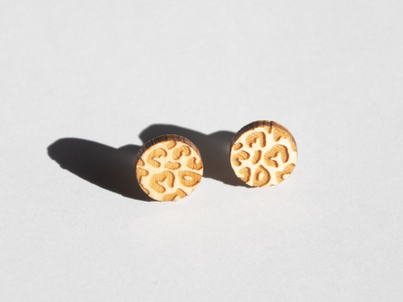 May include: A pair of wooden earrings with a leopard print design. The earrings are round and have a light brown color.
