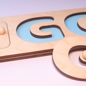 Op de afbeelding: Houten puzzel met het woord "GO". De puzzel toont de letters "G" en "O" uitgesneden uit lichtgekleurd hout, met een lichtblauwe achtergrond zichtbaar door de uitsparingen. Een kleine houten bal is inbegrepen.