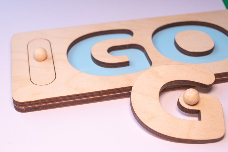 Op de afbeelding: Houten puzzel met het woord "GO". De puzzel toont de letters "G" en "O" uitgesneden uit lichtgekleurd hout, met een lichtblauwe achtergrond zichtbaar door de uitsparingen. Een kleine houten bal is inbegrepen.